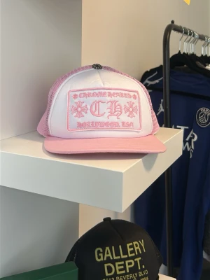  Chrome Hearts Rosa truckerkeps  - Snygg rosa och vit truckerkeps från Chrome Hearts med broderad logga och text på framsidan. Kepsen har mesh på sidorna och bak för extra ventilation och en böjd skärm. Perfekt accessoar för en streetwear-look.