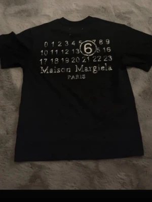 Maison Margiela svart t-shirt - Snygg svart t-shirt från Maison Margiela med ikoniskt tryck på ryggen med siffror och logga. Klassisk passform och rund hals. Tillverkad i mjuk bomull, perfekt för en stilren och trendig look.