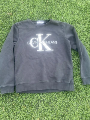 Svart sweatshirt från Calvin Klein - Svart sweatshirt från Calvin Klein Jeans med stort vitt CK-logotyptryck på bröstet. Tröjan har rund halsringning, ribbade muddar och är tillverkad i mjukt bomullsmaterial. Perfekt för en avslappnad och stilren look.