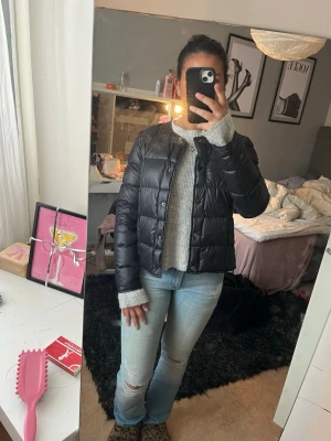 Vårjacka - Snygg svart dunväst med quiltad design och rund halsringning. Västen har tryckknappar framtill och en boxig, kortare passform som ger en trendig look. Perfekt att slänga över en stickad tröja för extra värme. Enkel och stilren utan synligt märke.