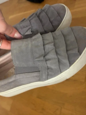 Dasia skor  med volangdetaljer - Snygga Dasia grå sneakers med coola volangdetaljer på ovandelen och vit sula. Skorna har slip-on-modell med elastiska partier på sidorna för enkel påtagning. Perfekta för dig som vill ha en unik touch på dina sneakers.