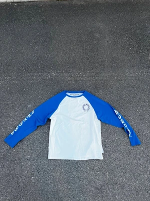 Blåvit långärmad topp Chrome Hearts - Säljer en blåvit långärmad topp från Chrome Hearts med blå ärmar och tryck på bröstet samt text på ärmarna. Klassisk basebollmodell med rund halsringning. Perfekt för dig som gillar streetwear och vill sticka ut.