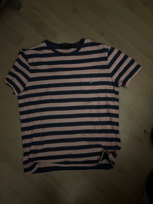 Randig t-shirt från Polo Ralph Lauren - Snygg t-shirt med breda rosa och marinblå ränder från Polo Ralph Lauren. Klassisk passform, kortärmad och med liten bröstficka med broderad logga. Perfekt för en avslappnad och trendig look.
