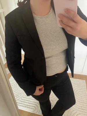Svart klassisk kavaj med två knappar - Snygg svart kavaj med figurnära passform och två knappar framtill. Kavajen har klassiska slag, två fickor med lock och en bröstficka. Perfekt för en stilren look. Materialet ser ut att vara en slät textil, troligen polyester eller bomullsblandning.