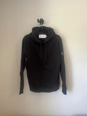 Svart hoodie från Calvin Klein Jeans - Snygg svart hoodie från Calvin Klein Jeans med stor CK-logga på vänster ärm och stor känguruficka framtill. Skick 8/10 