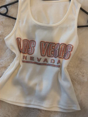 Vit ribbad linnetopp Las Vegas - Säljer en vit ribbad linnetopp med texten 'Las Vegas Nevada' i glittriga stenar framtill. Toppen är ärmlös och har en rund halsringning. Perfekt för dig som vill ha en cool och trendig look.