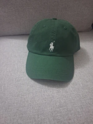 Grön keps från Polo Ralph Lauren - Snygg grön keps från Polo Ralph Lauren med klassisk vit broderad logga framtill och texten 'POLO' baktill. Justerbar rem bak för perfekt passform. Tillverkad i bomull och har en avslappnad, sportig look.