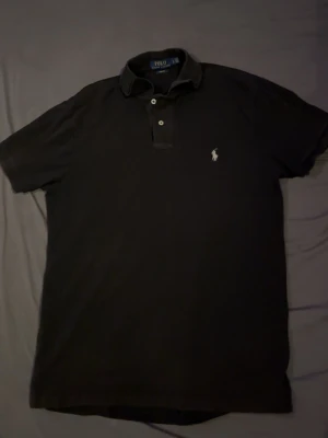 Svart pikétröja från Polo Ralph Lauren - En snygg svart pikétröja från Polo Ralph Lauren. Tröjan har krage, knappar framtill och är i slim fit-modell. Perfekt för en stilren och enkel look. Använt fåtal gånger.