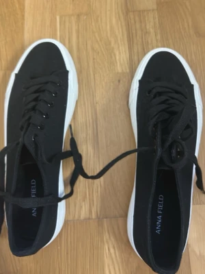 Svarta låga sneakers från Anna Field - Svarta sneakers från Anna Field med klassisk låg modell. Skorna har svart canvasmaterial, svarta skosnören och vit gummisula med vågigt mönster undertill. Perfekta för en stilren och avslappnad look.
