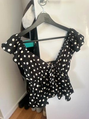 Prickig blus - Supersöt svart blus med vita prickar  . Blusen har korta volangärmar, v-ringning och knyt framtill. Perfekt för dig som gillar en lekfull och trendig stil. Luftigt material och figurnära passform.