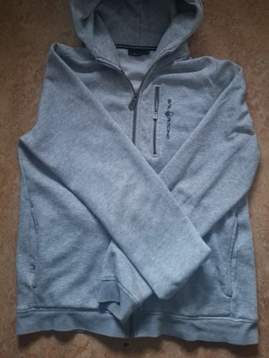 Ljusgrå hoodie från Sail Racing - Fet ljusgrå hoodie från Sail Racing med dragkedja framtill och broderad logga på bröstet. Hoodien har en bröstficka med dragkedja, två sidofickor och mjukt fleecefoder på insidan. Perfekt för chill dagar och streetwear-stil.