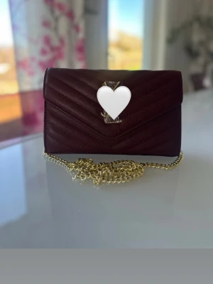Vinröd kuvertväska med kedja - Snygg vinröd kuvertväska med elegant guldmetallkedja. Väskan har klassisk V-formad frontflik och diskret guldig logga framtill. Perfekt och supergullig accessoar för att lyfta din outfit!💕 kan gå ned i pris vid snabb affär!! Mått: Längd: 22,5 cm, höjd: 15 cm, djup: 5 cm