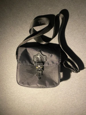 Svart axelväska från Trapstar - Snygg svart axelväska från Trapstar med justerbar axelrem och coolt spänne framtill. Väskan har en fyrkantig form och är gjord i slitstarkt tyg med detaljer i mesh och metall. Perfekt för att bära det viktigaste och ger en streetwear vibe.