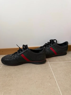 Svarta Gucci sneakers med röd/grön rand - Snygga svarta sneakers från Gucci med klassiska gröna och röda ränder på sidan. Skorna har lågt skaft, svarta skosnören och präglat GG-mönster i lädret. Perfekta för dig som vill ha en exklusiv och stilren look.