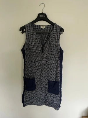 Mönstrad blå ärmlös byxdress - Snygg ärmlös byxdress i mörkblått med småvitt mönster framtill och helblå baksida. Två stora fickor framtill i matchande blått. Lätt och luftig modell, perfekt för varma dagar. Ingen synlig dragkedja eller knappar.