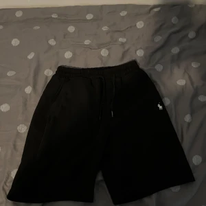 Svarta shorts från Polo Ralph Lauren - Svarta shorts från Polo Ralph Lauren med klassisk vit broderad logga på benet. Modellen har elastisk midja med snörning och två sidofickor. 14-16