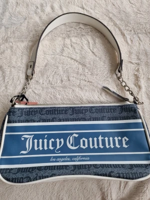 Juicy Couture blå axelväska - Snygg axelväska från Juicy Couture i blått och vitt med tydlig logga framtill. Väskan har ett brett vitt axelband med kedjedetaljer och dragkedja upptill. Materialet är en mix av textil och konstläder, perfekt för dig som gillar streetstyle och loggor.