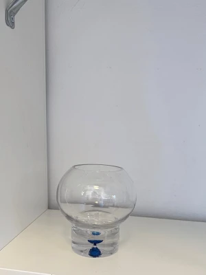 Glasvas med blå detalj - Säljer en rund glasvas med en unik blå detalj i botten. Vasen är genomskinlig och har en bred öppning upptill, perfekt för blommor eller dekoration. Tillverkad i glas och har en modern, stilren design.
