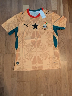 Ghana fotbollströja Puma GUL. Storlek M - Snygg beige Ghana fotbollströja från Puma med svarta detaljer, svart stjärna på bröstet och landslagsemblem. Tröjan har mönstrade partier, gröna inslag på sidorna och röd krage. Perfekt för fotboll eller träning.