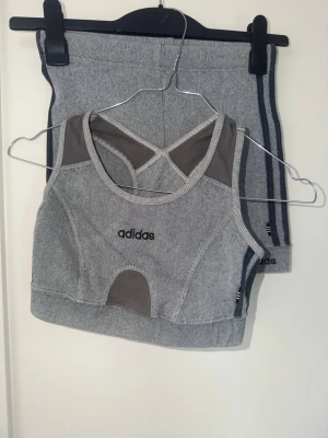 Grått set från Adidas med sportig look - Snyggt grått set från Adidas med sport-bh och matchande shorts. Setet har mörkgrå detaljer, broderad Adidas-logga framtill och är gjort i mjukt, stretchigt material. Perfekt för dig som gillar sportig och bekväm stil.