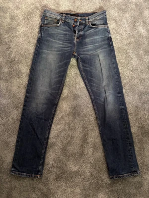 Blå jeans från Nudie - Snygga blåa jeans från Nudie med klassisk femficksdesign och orangea sömmar. Jeansen har en lätt tvättad look med slitningar på låren och bakfickor med vågig brodyr. Perfekta för en avslappnad och trendig stil. Jeansen är i bra skick. Hör av er vid minsta fundering!