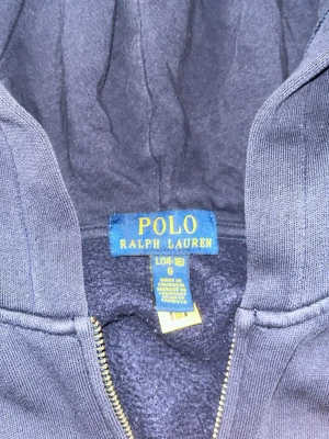 Mörkblå hoodie från Polo Ralph Lauren - Snygg mörkblå hoodie från Polo Ralph Lauren med dragkedja och klassisk röd logga broderad på bröstet. Hoodien har huva och är tillverkad i mjukt bomullsmaterial som känns skönt mot huden. Perfekt för en avslappnad och stilren look. Litet tecken på slitage