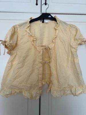 Gul romantisk blus med rosetter - Söt ljusgul blus med korta puffärmar och volangkant. Blusen har knytband framtill och spetsdetaljer längs krage och nederkant. Perfekt för en romantisk look och tillverkad i ett lätt tyg som känns luftigt. 