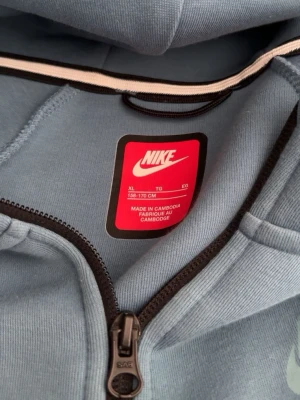 Nike blå och vit hoodie med dragkedja - Snygg hoodie från Nike i blått och vitt med hel dragkedja framtill. Hoodien har huva och svarta detaljer längs dragkedjan och på ärmen. Perfekt för en sportig och avslappnad stil. Tillverkad i mjukt material för extra komfort.