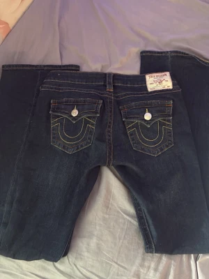 Mörkblå True Religion jeans - Snygga mörkblå jeans från True Religion med ikoniska bakfickor och kontrastsömmar. Klassisk femficksmodell med silverfärgade knappar och tydlig logopatch bak. Perfekta för dig som gillar streetwear och vill sticka ut med detaljer.