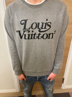 Grå sweatshirt från Louis Vuitton - Snygg grå sweatshirt från Louis Vuitton med stor svart logga på bröstet. Tröjan har rund halsringning, långa ärmar och ribbade muddar vid ärmslut och nederkant. Perfekt för en avslappnad och trendig look. Det står XXL men sitter som en M-L