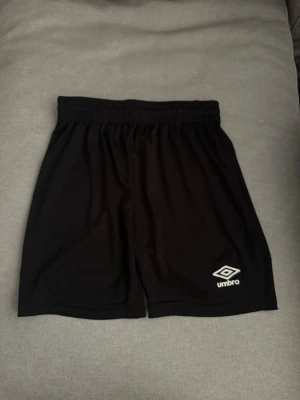 Svarta träningsshorts från Umbro - Svarta träningsshorts från Umbro med elastisk midja och logga på både fram- och baksidan. Tillverkade i ett lätt och bekvämt syntetmaterial som passar perfekt för sport och aktivitet.
