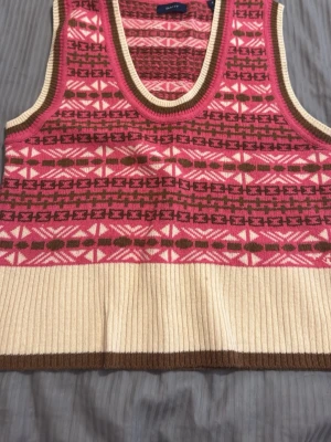 Mönstrad stickad väst från GANT - Cool stickad väst från GANT i storlek S med rosa, bruna och vita geometriska mönster. Västen har bred ribbad beige nederkant och bruna detaljer vid ärm och halsringning. Perfekt att styla över en skjorta eller t-shirt för en trendig look.