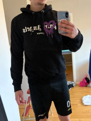 Svart Palm Angels hoodie med lila hjärta - Svart hoodie från Palm Angels med vit text och ett lila spraymålat hjärta på bröstet. Klassisk passform med huva och dragsko, samt ribbade muddar. Trycket på framsidan ger en streetwear vibe. Perfekt för dig som gillar trendiga och unika plagg. Modell på bild är 188 och väger 76kg