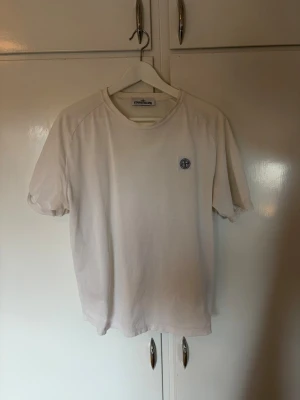 Vit t-shirt från Stone Island - Säljer en clean vit t-shirt från Stone Island med klassisk logga på bröstet. T-shirten har rund hals och korta ärmar, perfekt basic-plagg till garderoben. Tillverkad i mjuk bomull för skön känsla hela dagen.