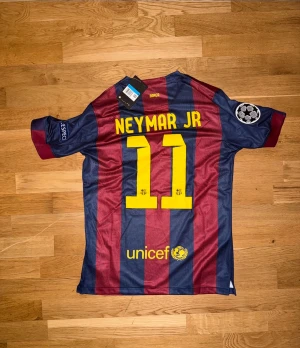 Barcelona Neymar Jr matchtröja retro - Säljer en FC Barcelona matchtröja från Nike med Neymar Jr och nummer 11 på ryggen. Tröjan har blå och vinröda ränder, gula detaljer och officiella klubbmärken. Materialet är lätt och andas, perfekt för fotboll eller träning.