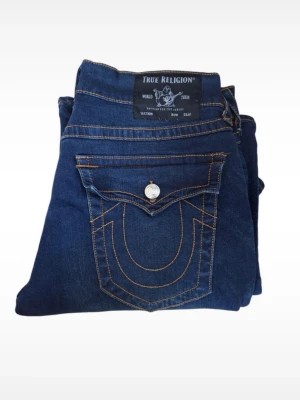True Religion mörkblå jeans - Mörkblå jeans från True Religion med klassiska kontrastsömmar och stora bakfickor med lock och silverfärgade knappar. Jeansen har en rak passform och är tillverkade i denim. Perfekta för dig som gillar streetwear och snygga detaljer.