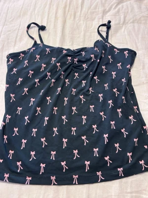  linnetopp med rosa rosetter - Söt linnetopp med tunna axelband som knyts upptill. Toppen har ett gulligt mönster av små rosa rosetter över hela tyget och en lätt rynkning vid bysten. Perfekt för en trendig och lekfull look.