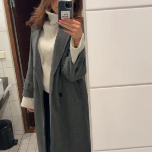 Lång grå kappa med knappar - Säljer min långa fina gråa kappa från Nelly💕 jag är 170 cm lång och den sitter perfekt på mig och går ner till backen😍 perfekt nu till våren, den är fodrad och varm. Se bild tre för hur den är i materialet lite närmare😊