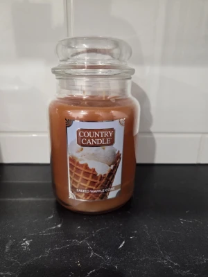 Salted Waffle Cone 680g - Säljer mitt Country Candle ljus som är oanvänt. Doftljuset Country Candle Salted Waffle Cone har en söt gourmanddoft med toner av smörigt våffelrån, regnbågsströssel, salt karamellsås och vaniljglass. Doftprofil och egenskaper Doften beskrivs som en härlig sommarkänsla som för tankarna till ett glasskafé.  Doftnoter: Smörigt våffelrån, regnbågsströssel, salt karamellsås och vaniljglass. Doftkaraktär: Söt, vaniljig och gourmand. Egenskaper: Ljuset har en hög doftkoncentration (27-50% mer doftolja