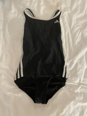 Svart baddräkt från Adidas - Snygg svart baddräkt från Adidas med vita detaljer och klassiska ränder på sidorna. Modellen har smala axelband och en sportig rygg med öppning. Perfekt för simträning eller strandhäng. Materialet är snabbtorkande och stretchigt för bästa komfort.❤️