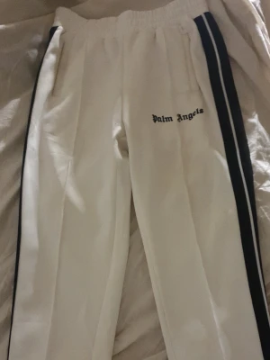 Vita mjukisbyxor - Vita mjukisbyxor med svarta sidoremsor och logotyp på låret. Byxorna har elastisk midja och raka ben, perfekt för en avslappnad och trendig look. Materialet är mjukt och bekvämt. A kopia 