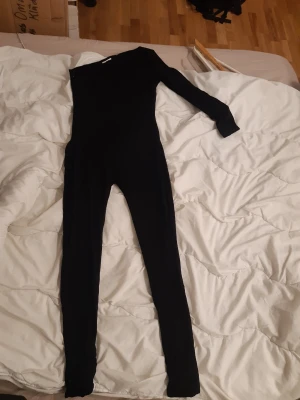 Svart one shoulder jumpsuit från Bodssy  - Säljer en svart, långärmad jumpsuit med one shoulder-design. Tight passform och långa ben ger en snygg siluett. Perfekt för dig som vill sticka ut med en stilren och trendig look. Materialet: Modal och elastane,känns stretchigt och bekvämt. Dubbelsydd. Skicket är helt nytt.