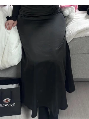 Svart långkjol i satin material - En svart lång satinkjol med figurnära passform och lätt utsvängd nederdel. Kjolen är enkel och stilren, perfekt att matcha med olika toppar.