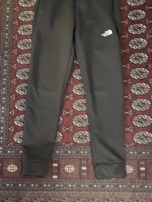 Svarta mjukisbyxor från The North Face - Säljer ett par svarta mjukisbyxor från The North Face med logga på vänster ben. Byxorna har smal passform, ribbade muddar vid benslut och är gjorda i ett mjukt material som känns skönt mot huden. Perfekta för chill eller träning.