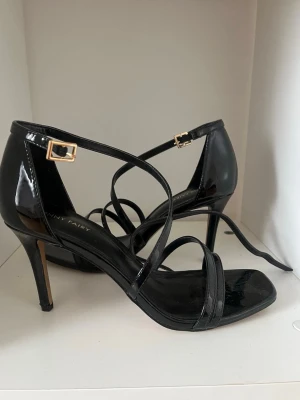 Svarta lackade sandaletter med klack - Säljer ett par svarta sandaletter från Penny Fairy i glansigt lackmaterial. De har smala remmar över foten, öppen tå och en elegant klack. Spänne i guld vid ankeln och fyrkantig tå ger en trendig look. Perfekta för att lyfta vilken outfit som helst.