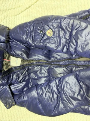 Blå dunjacka från Moncler - Snygg blå dunjacka från Moncler med glansig finish och klassisk logotyp på bröstfickan. Jackan har dragkedja, tryckknappar och en innerficka med serietidningsmönster. Perfekt för kalla vinterdagar och riktigt varm tack vare dunfyllningen.