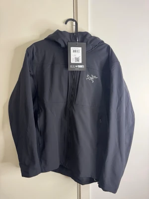 Svart vindjacka från Arc'teryx GAMMA MX HOODY - Snygg svart vindjacka från Arc'teryx med huva och dragkedja framtill. Jackan har två sidofickor med dragkedja och en stilren logga på bröstet. Tillverkad i ett lätt och slitstarkt material som passar perfekt för blåsiga dagar.