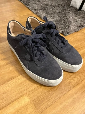 Mörkblå mockasneakers med vita sulor - Snygga mörkblå sneakers i mocka med klassisk låg profil och vita gummisulor. Skorna har matchande mörkblå skosnören och en enkel, stilren design som passar till det mesta. Insidan är ljusbeige och ger en schysst kontrast till utsidan.