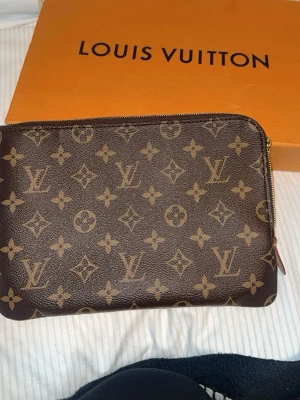 Louis Vuitton Monogram Kuvertväska - Säljer en klassisk brun kuvertväska från Louis Vuitton med ikoniskt monogrammönster i beige. Väskan har en smidig dragkedja upptill och är tillverkad i tåligt canvasmaterial. Perfekt för dig som vill ha en lyxig accessoar med tidlös design.