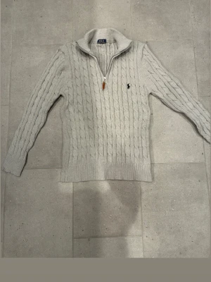  kabelstickad tröja Polo Ralph Lauren - Kabelstickad ljusgrå tröja från Polo Ralph Lauren med halv dragkedja och hög krage. Tröjan har det klassiska broderade logotypen på bröstet och ribbade muddar. Perfekt för dig som gillar stilren och tidlös design.